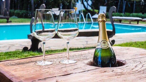 A wooden table with two empty wine glasses and a bottle of champagne in front of a swimming pool.
