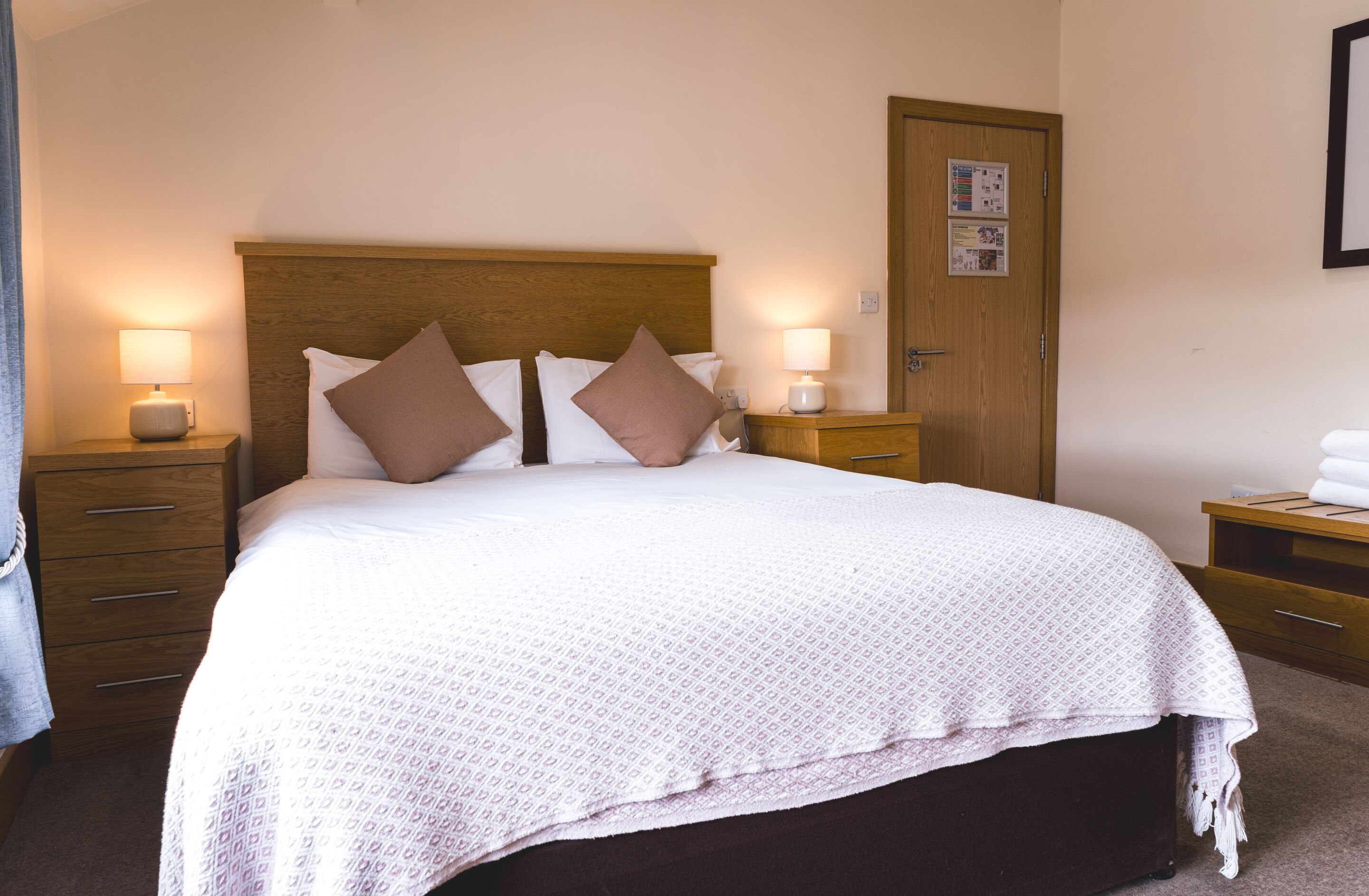 A hotel room with a large bed, two bedside tables with lamps, a wooden headboard, a wooden door, and a small wooden bench.
