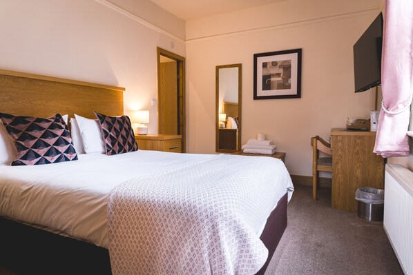 A hotel room with a bed, bedside table, lamp, mirror, framed picture, and a desk with a chair. - Horse and Farrier (Otley)