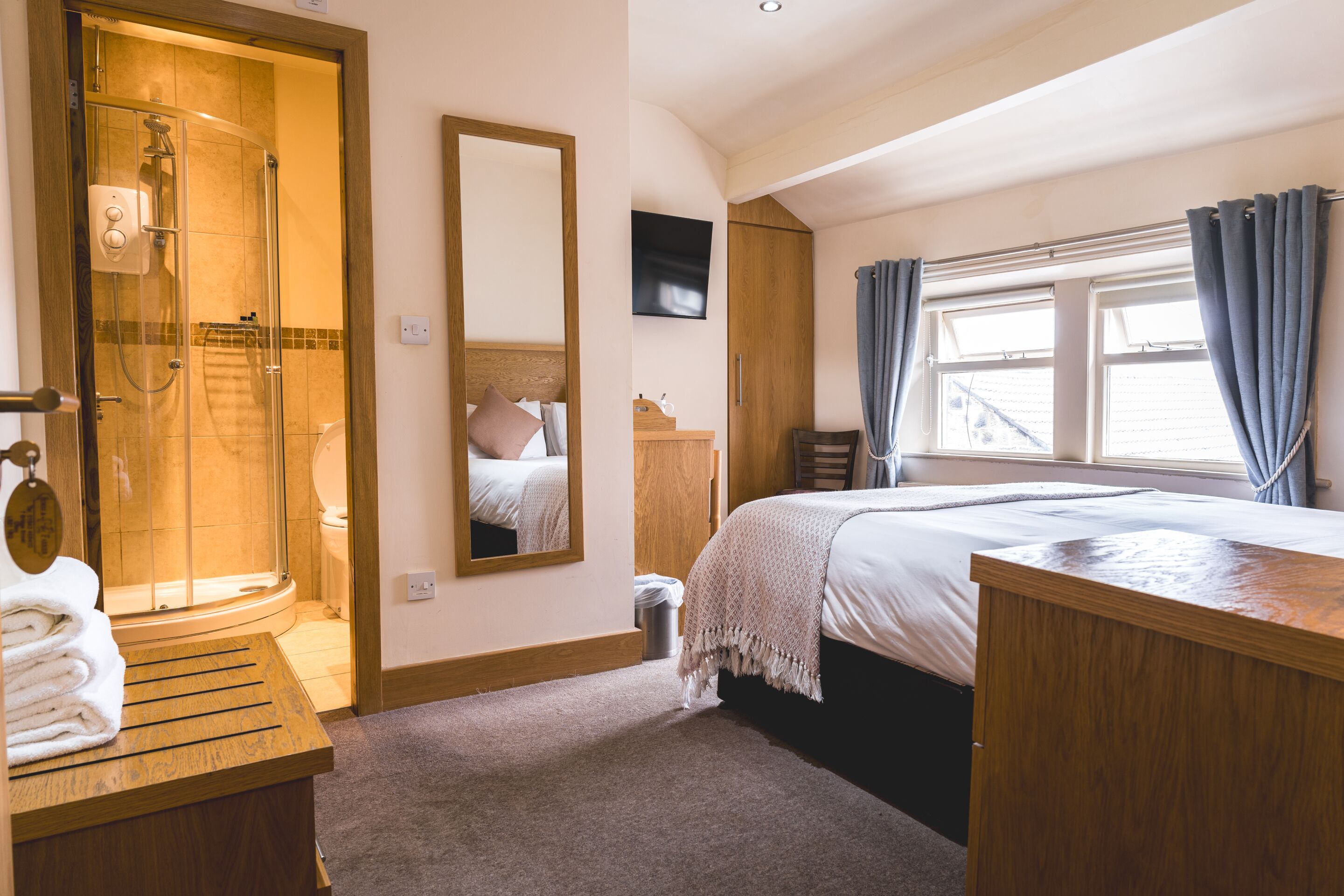 A hotel room with a shower, a bed, a wooden desk, a mirror, and a TV.