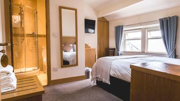 A hotel room with a shower, a bed, a wooden desk, a mirror, and a TV.