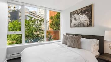 Une chambre avec une grande fenêtre, un lit, une table de chevet, une lampe et un tableau encadré au mur.