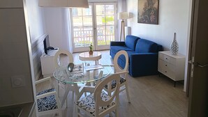 Uma sala de estar moderna com um sofá azul, uma mesa de jantar de vidro com cadeiras, um abajur branco e um quadro na parede.