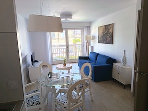 A modern living room with a blue sofa, a glass dining table with chairs, a white lamp, and a painting on the wall.