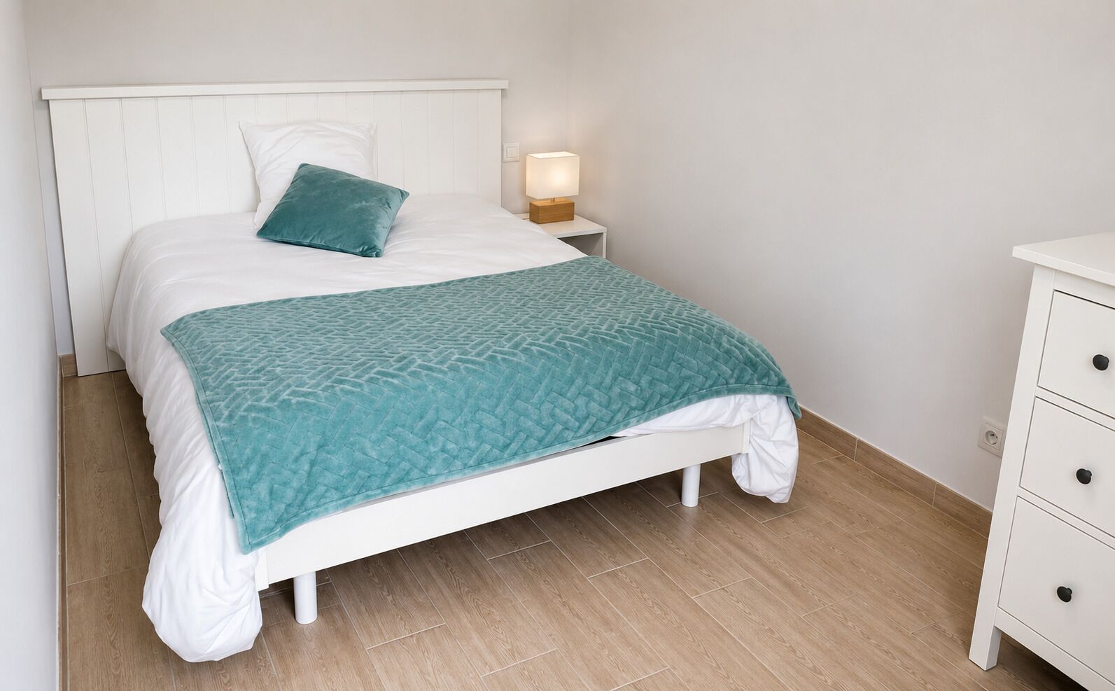 A bed with a white headboard, a teal throw blanket, and a bedside lamp.