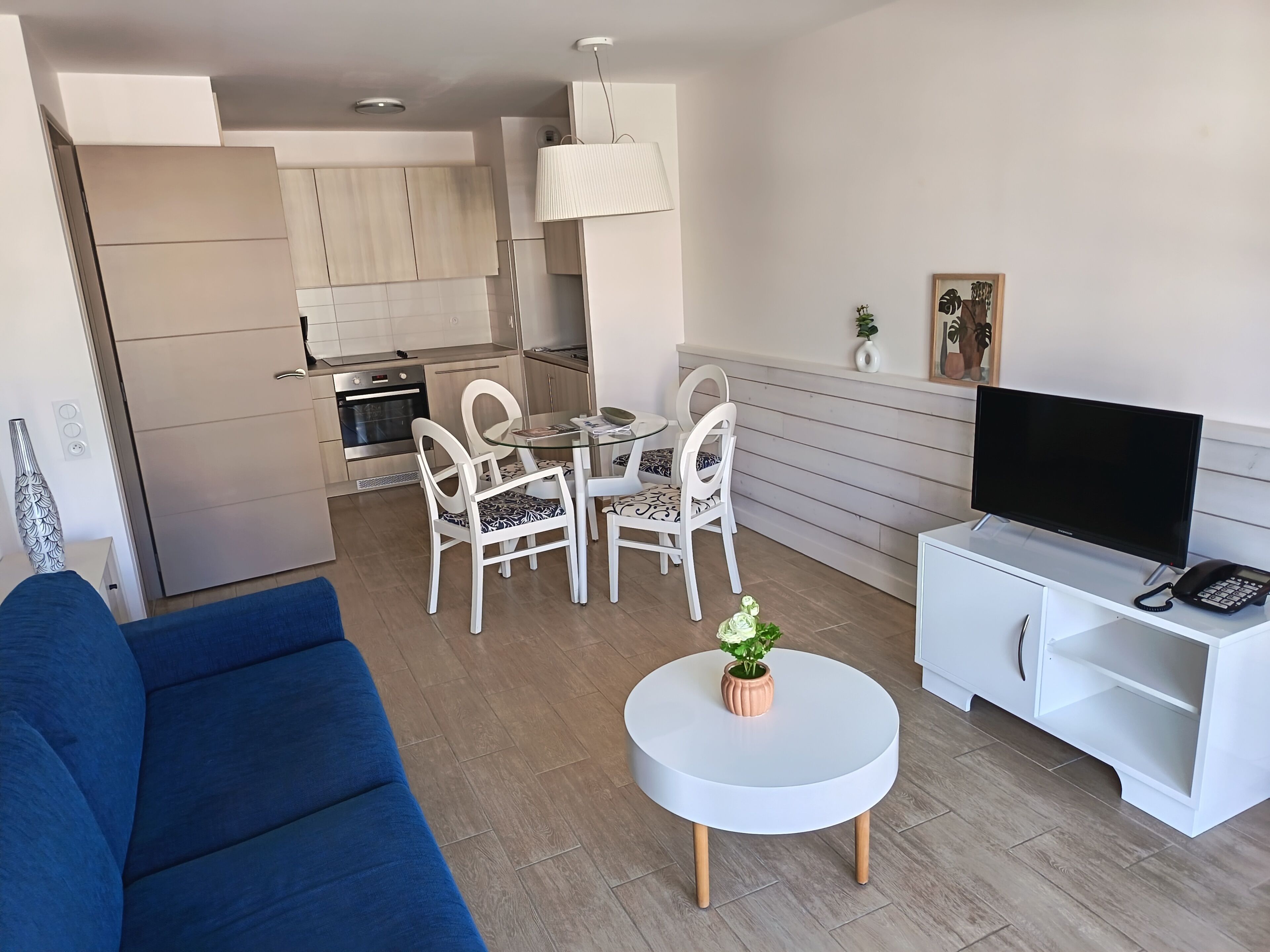 Ein modernes Wohnzimmer mit einem blauen Sofa, einem runden Couchtisch mit Topfpflanze, einem Essbereich mit weißen Stühlen und einer Küche im Hintergrund.