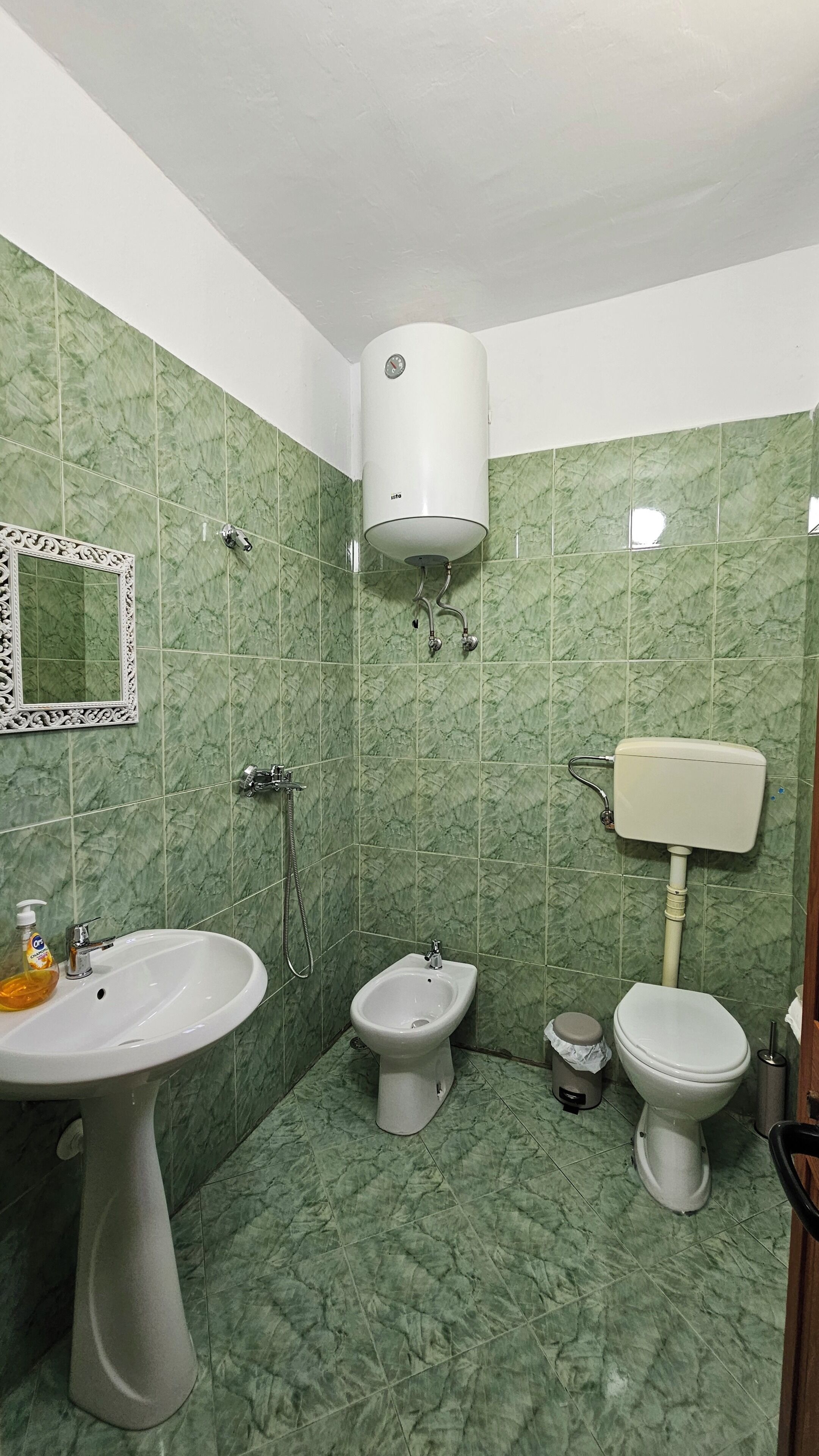 Un baño con baldosas de mármol verde, un lavamanos pedestal, un bidé, un inodoro y un calentador de pared.