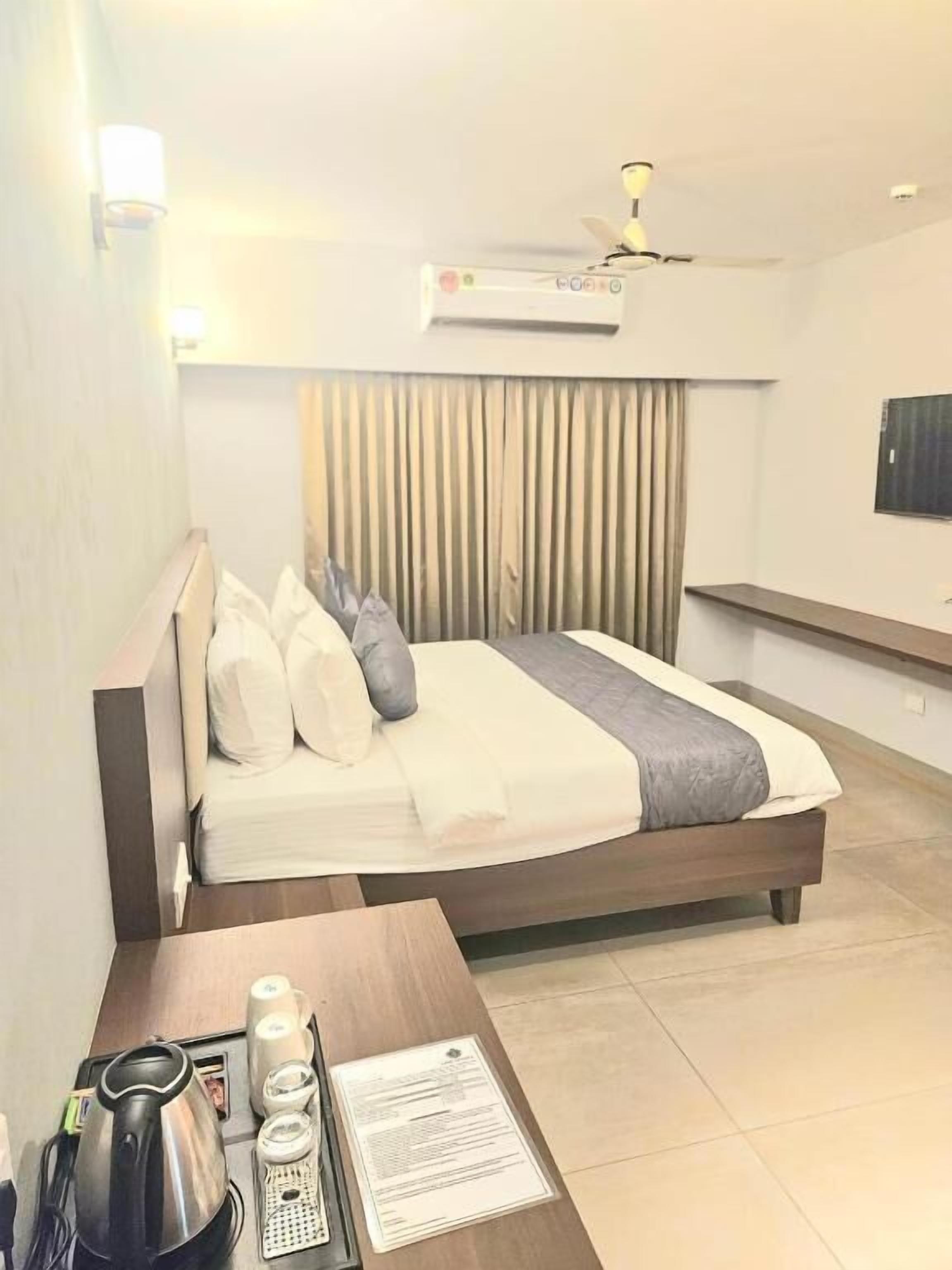 A hotel room with a bed, bedside table with a kettle, cups, and a menu, an air conditioning unit, and a ceiling fan.