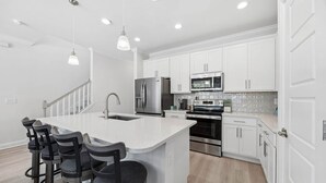A modern kitchen with white cabinets, stainless steel appliances, a central island with bar stools, and pendant lighting. - Beautiful 3BR 3BA Townhome~ Steps from the Beach! (Fernandina Beach)