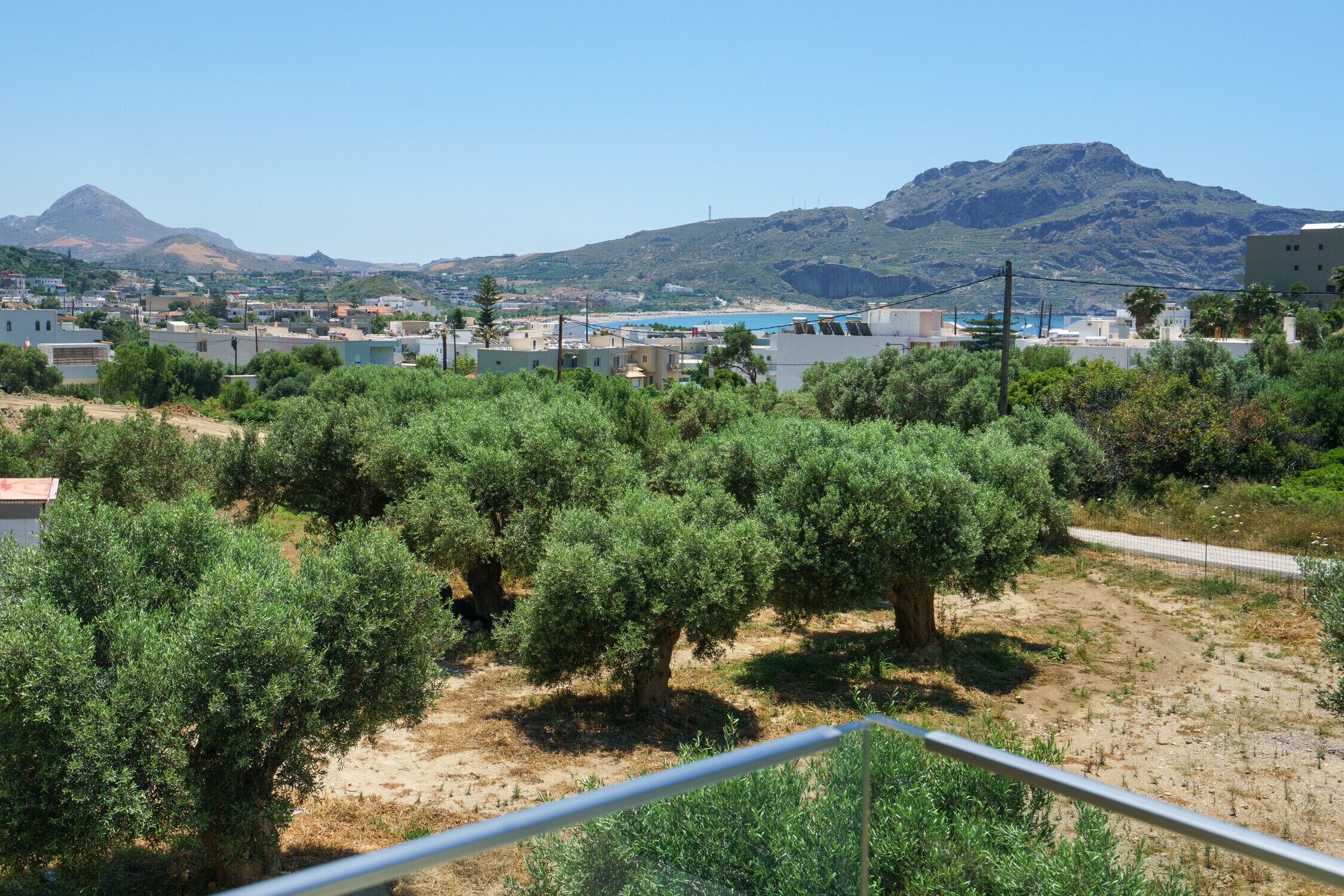 Vista de un pueblo costero con olivares, edificios y montañas al fondo.
