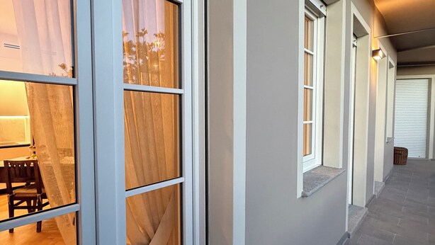 Un pasillo con una puerta de madera, un ventanal con vista a una habitación y un suelo de baldosas.