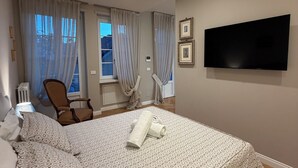 Un dormitorio con cama, una silla, un televisor y un ventanal con cortinas.