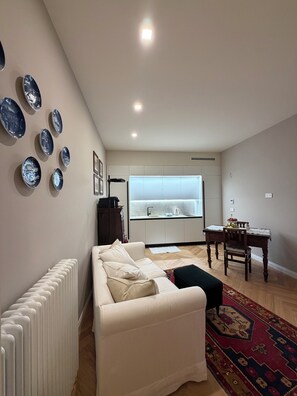 A modern living room with a white sofa, a dining table with chairs, a kitchen in the background, and decorative plates on the wall. - Cozy apartment for 2 people in the heart of Alba. (Alba)