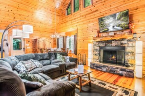 A log cabin interior with a fireplace, a sofa, a dining area, and a television mounted above the fireplace. - Kells Creek Cabin (Ellijay)