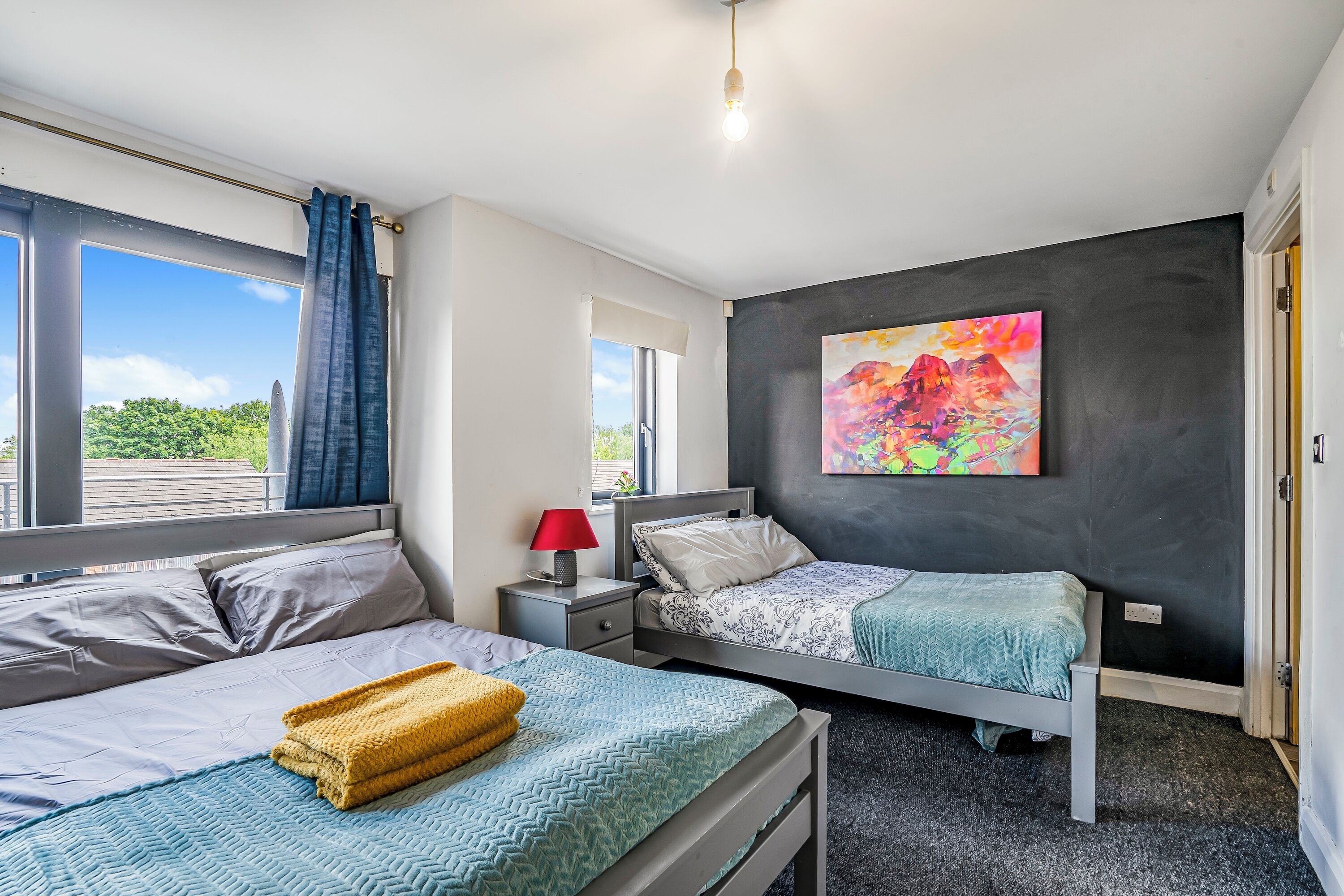 A bedroom with two beds, a nightstand, a lamp, a painting, and a window with curtains.