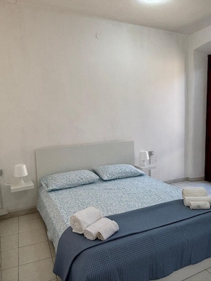 A bedroom with a bed, two pillows, a nightstand with a lamp, and rolled towels.