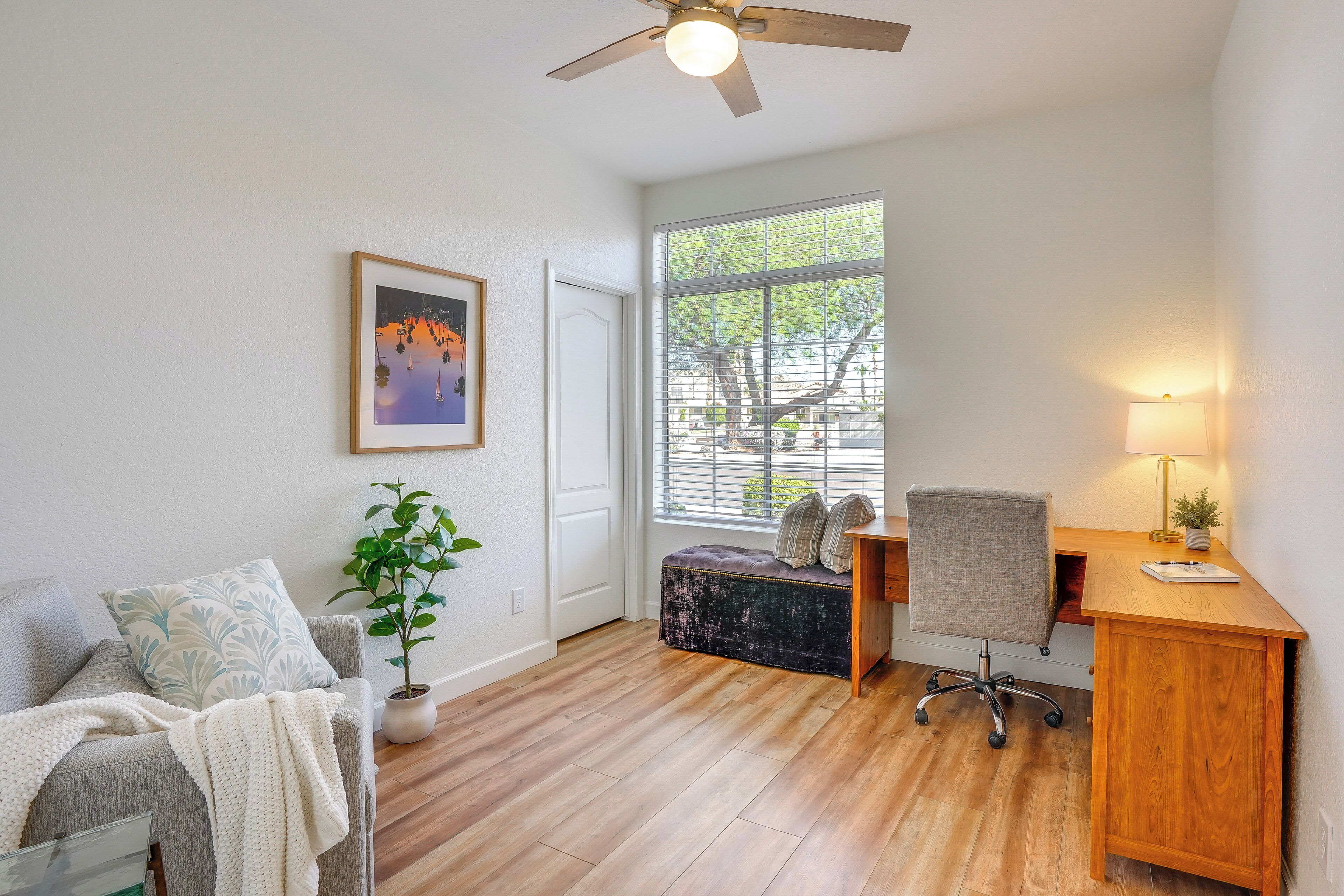 Una habitación con piso de madera, un escritorio con una silla, un sofá con una manta, un ventilador de techo, una ventana con persianas y un cuadro en la pared.