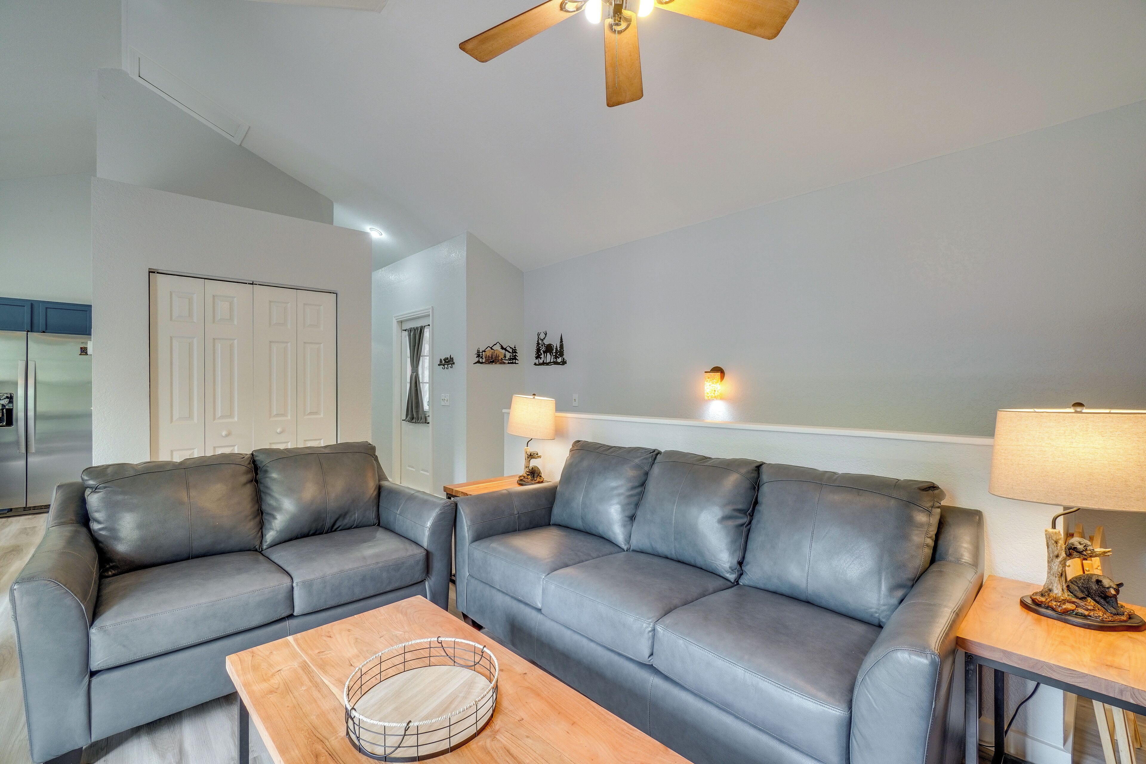 A living room with two grey leather sofas, a wooden coffee table, a ceiling fan, a wall-mounted lamp, and a door with a coat closet.