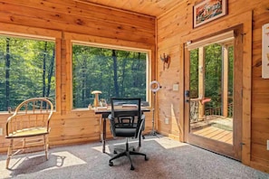 Interior de una cabaña de madera con un escritorio, dos sillas, ventanas amplias y una puerta que da al exterior.