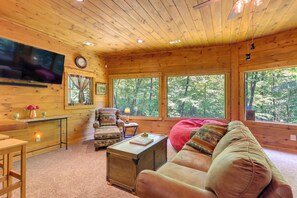 Ein gemütliches Blockhaus-Interieur mit Fernseher, Holzwänden, großen Fenstern, einer Couch, einem Sessel und einem kleinen Tisch.