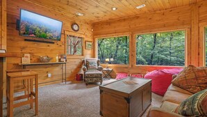 A wooden cabin interior with a television, a sofa, a coffee table, a chair, and a window with a forest view. - Forest Sanctuary, a Lakeside Retreat (Blue Ridge)