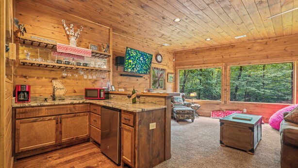 A wooden cabin interior with a kitchen, dining area, and large windows overlooking a forest. - Forest Sanctuary, a Lakeside Retreat (Blue Ridge)