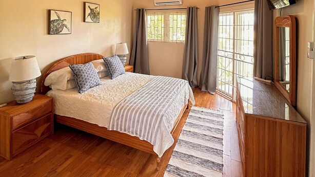 A bedroom with a wooden ceiling, a bed with a striped bedspread, a bedside table with a lamp, a TV mounted on the wall, and a window with curtains. - Spacious  2 Separate Units apartment Sleeps 8 in beautiful Rodney Bay with AC (Rodney Bay)