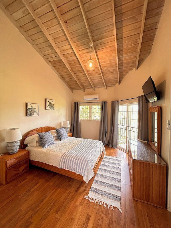 A bedroom with a wooden ceiling, a bed with a striped bedspread, a bedside table with a lamp, a TV mounted on the wall, and a window with curtains. - Spacious  2 Separate Units apartment Sleeps 8 in beautiful Rodney Bay with AC (Rodney Bay)