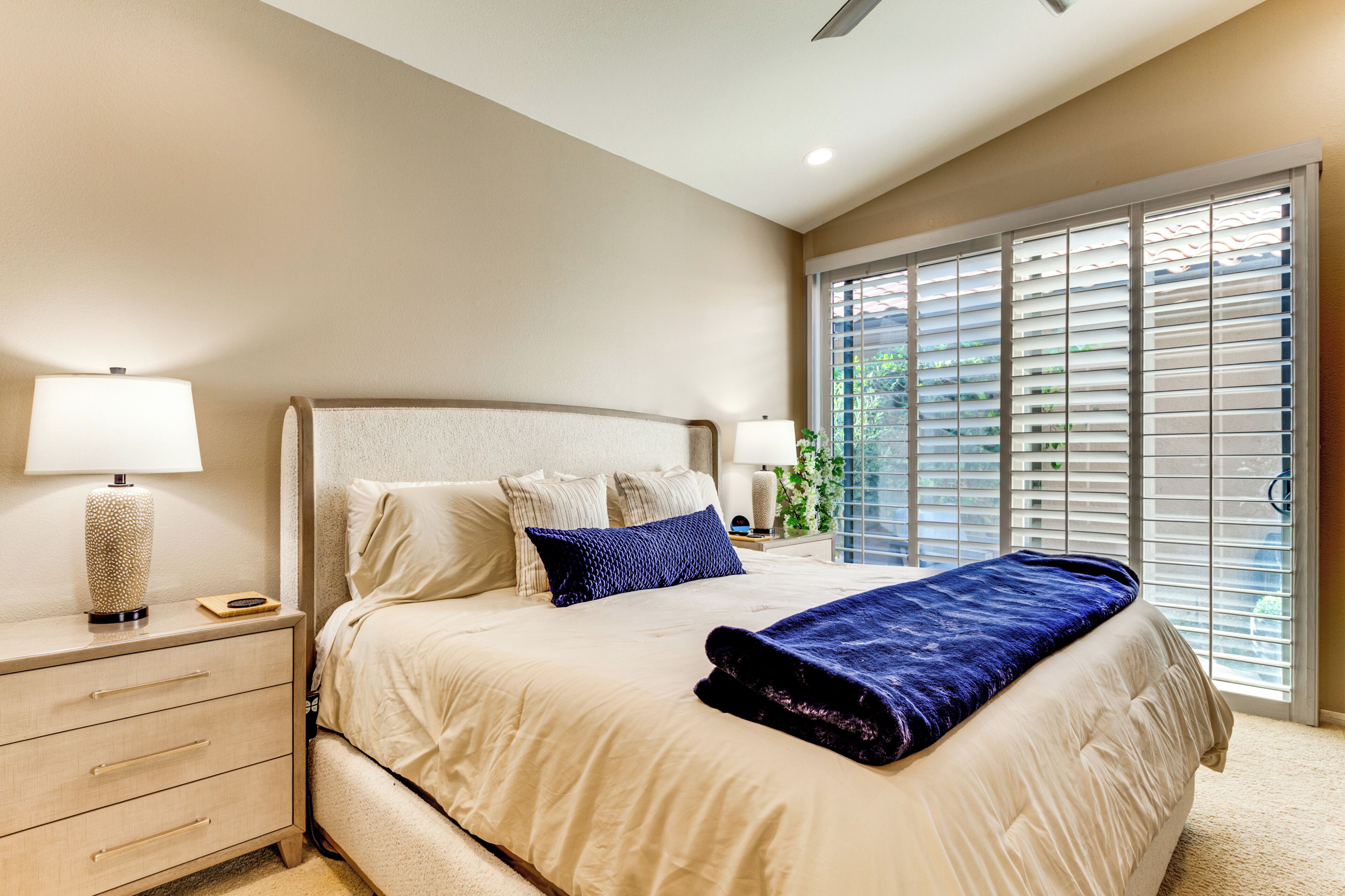 A bedroom with a bed, nightstands, lamps, and a large window with blinds.