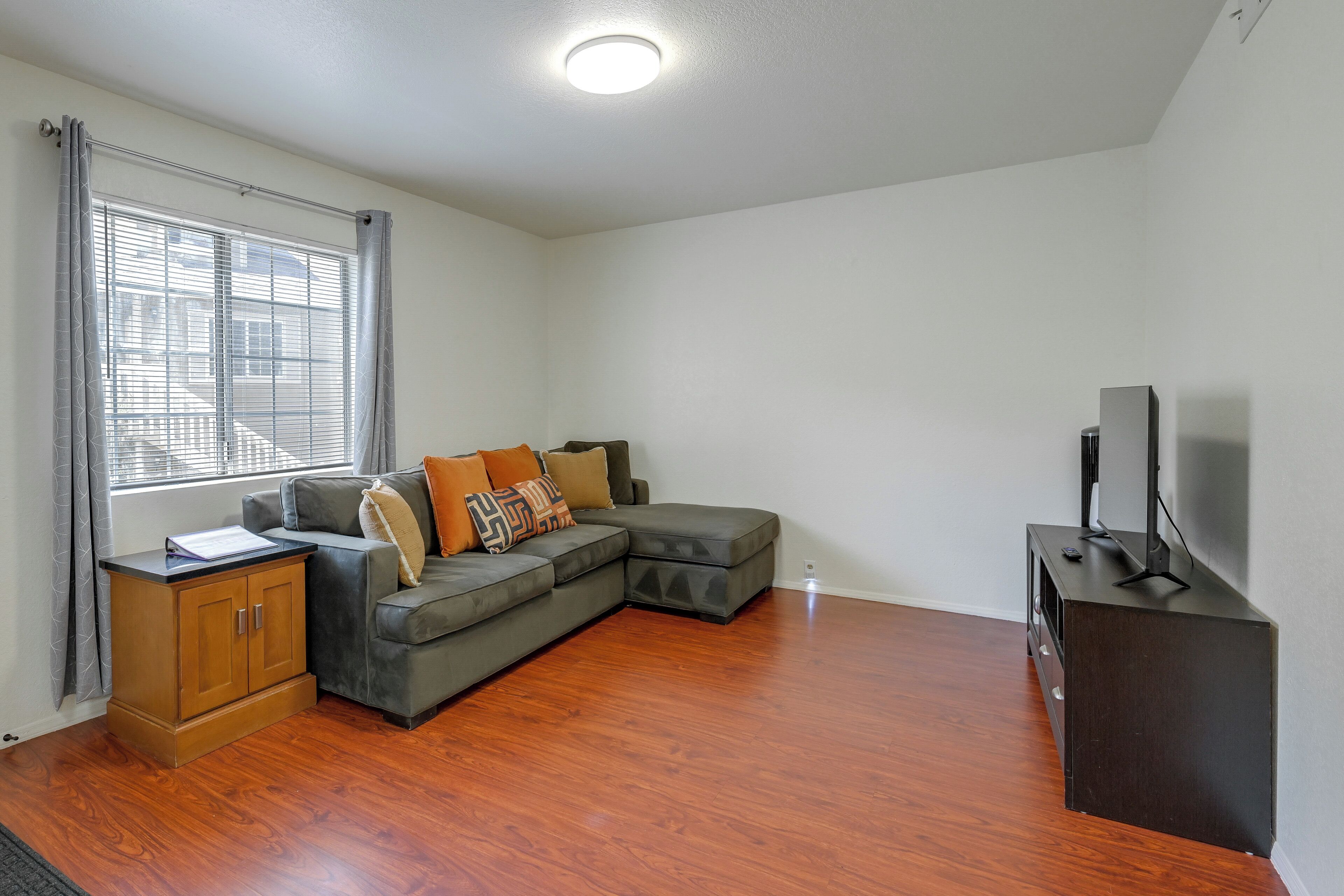 A living room with a sectional sofa, a wooden cabinet, a television on a stand, and a window with blinds.