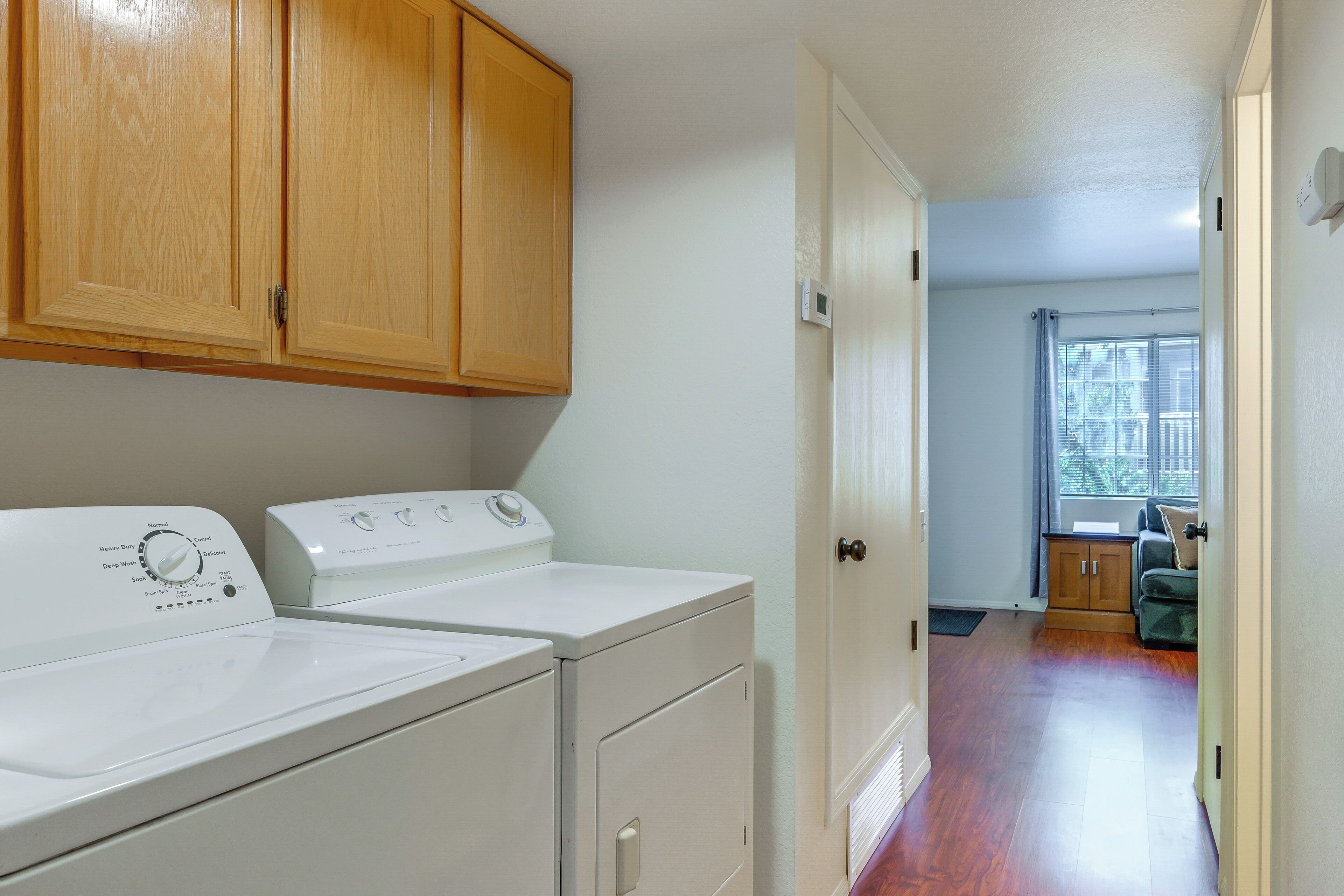 A laundry room with a washer and dryer, wooden cabinets, and a view into a living area through an open door.
