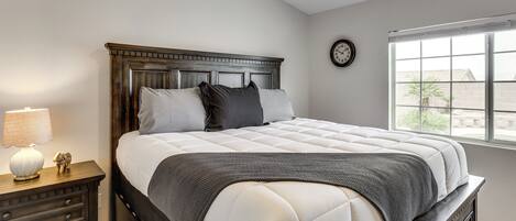 Quarto com cama, mesa de cabeceira, abajur, relógio e janela.