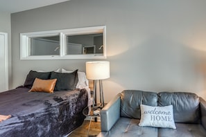 A bedroom with a bed, a bedside lamp, a sofa with a 'welcome home' pillow, and a large mirror above the bed.