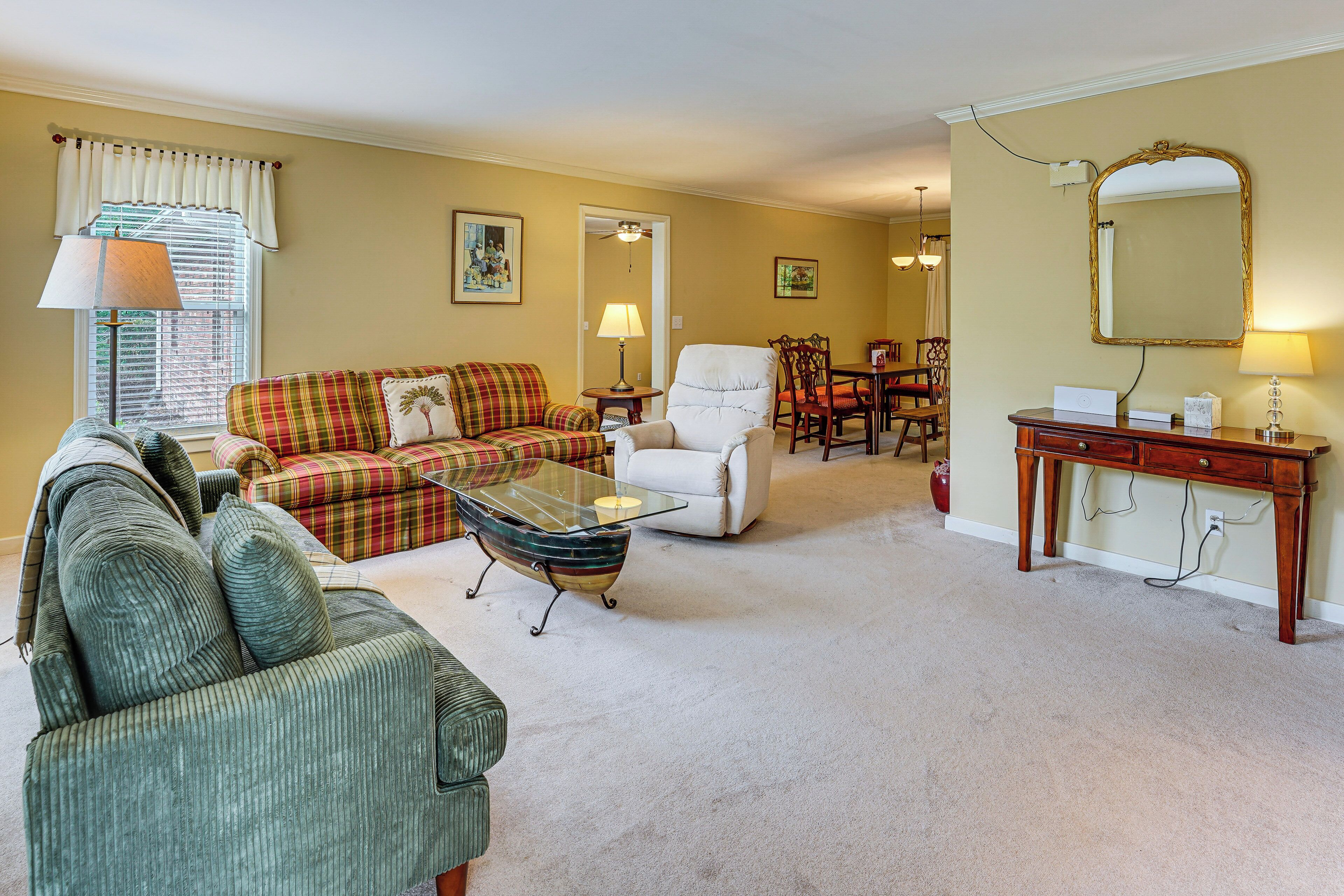 A living room with a green sofa, a plaid sofa, a glass coffee table, a white armchair, and a dining area with a table and chairs.