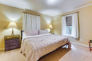 Quarto com cama, duas mesas de cabeceira, penteadeira e duas janelas com cortinas.