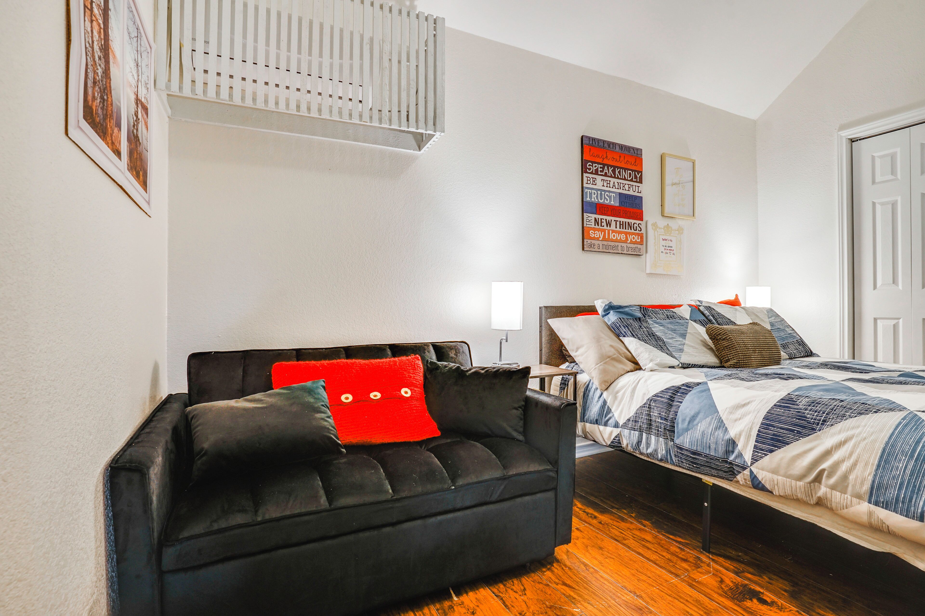 A bedroom with a bed, a black sofa with red cushions, a white wall, and a door.