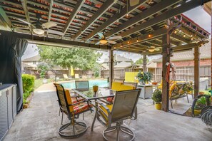 Een terras buiten met een glazen tafel, metalen stoelen, een plafondventilator en een zwembad op de achtergrond.