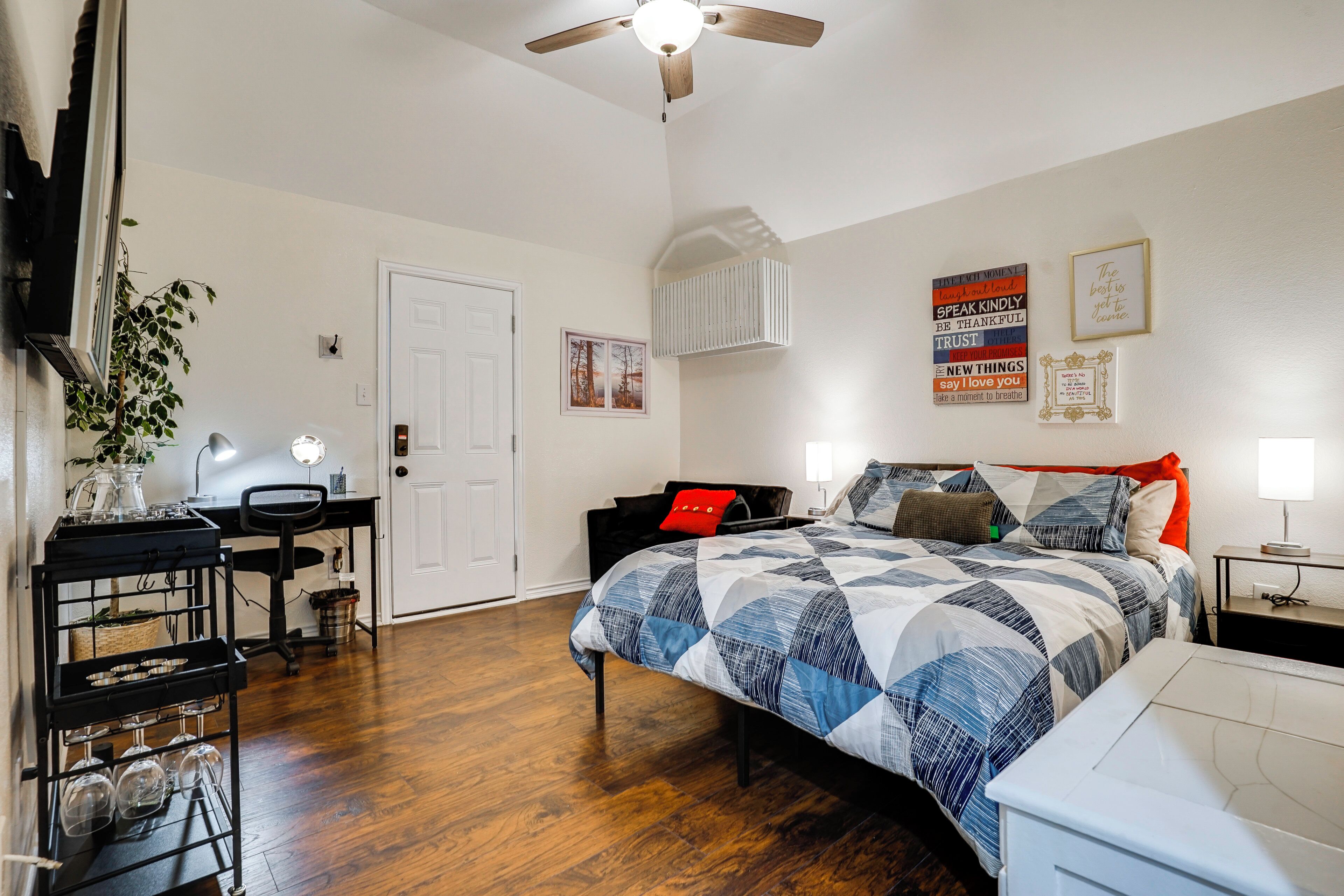A bedroom with a bed, a desk, a chair, a cart with glasses, and a ceiling fan.