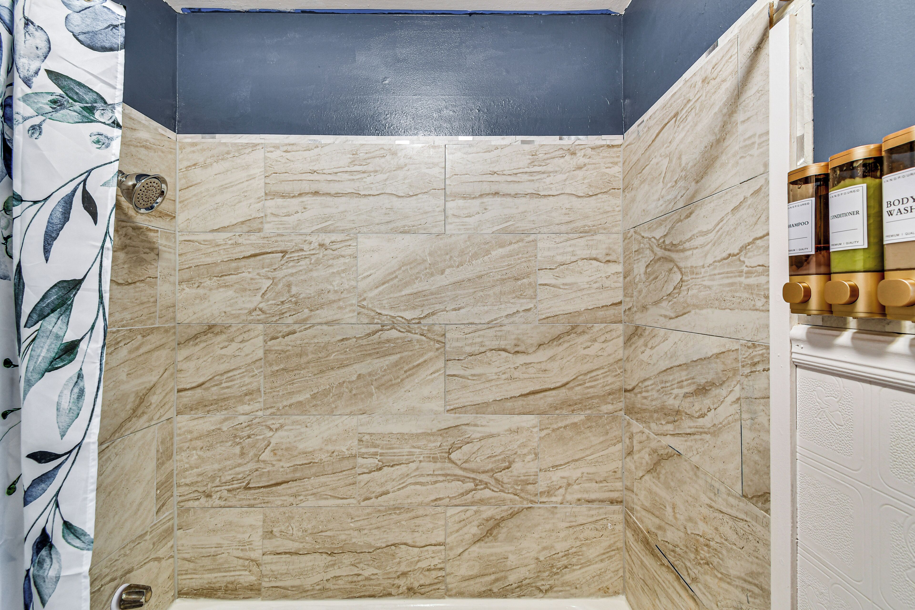 A shower with beige marble tiles, a blue wall, and a shower curtain with a leaf pattern.