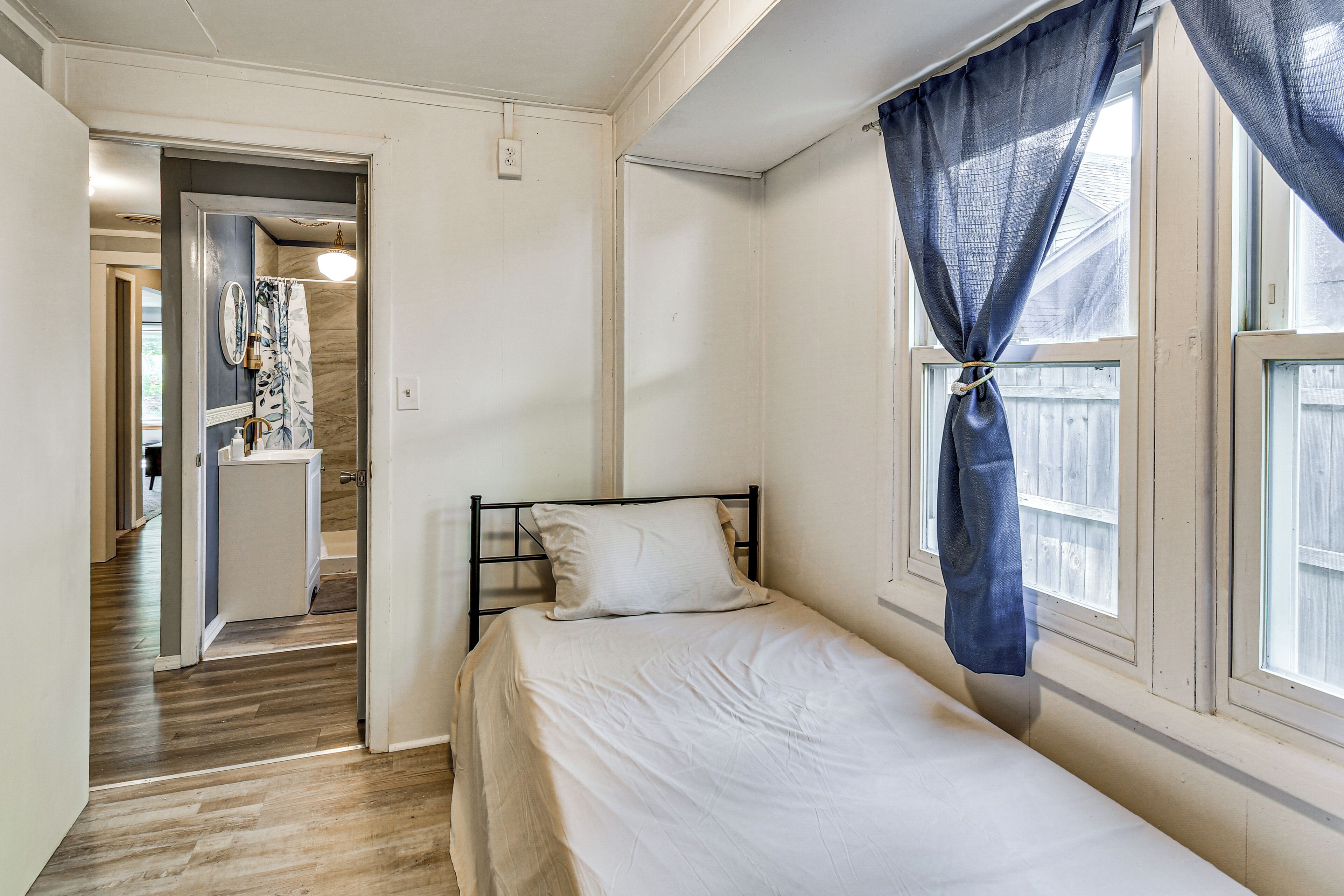 Ein Schlafzimmer mit einem Einzelbett, einem Fenster mit Vorhängen, einer Tür zu einem Badezimmer und einem Flur.