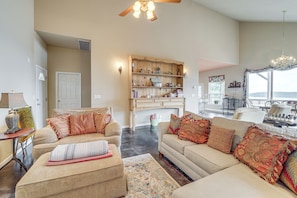 A spacious living room with a large sofa, a coffee table, and a wooden bookshelf. There is a dining area with a table and chairs visible in the background. - Sunset View Deck! Lake Eufaula Family Getaway (Stigler)