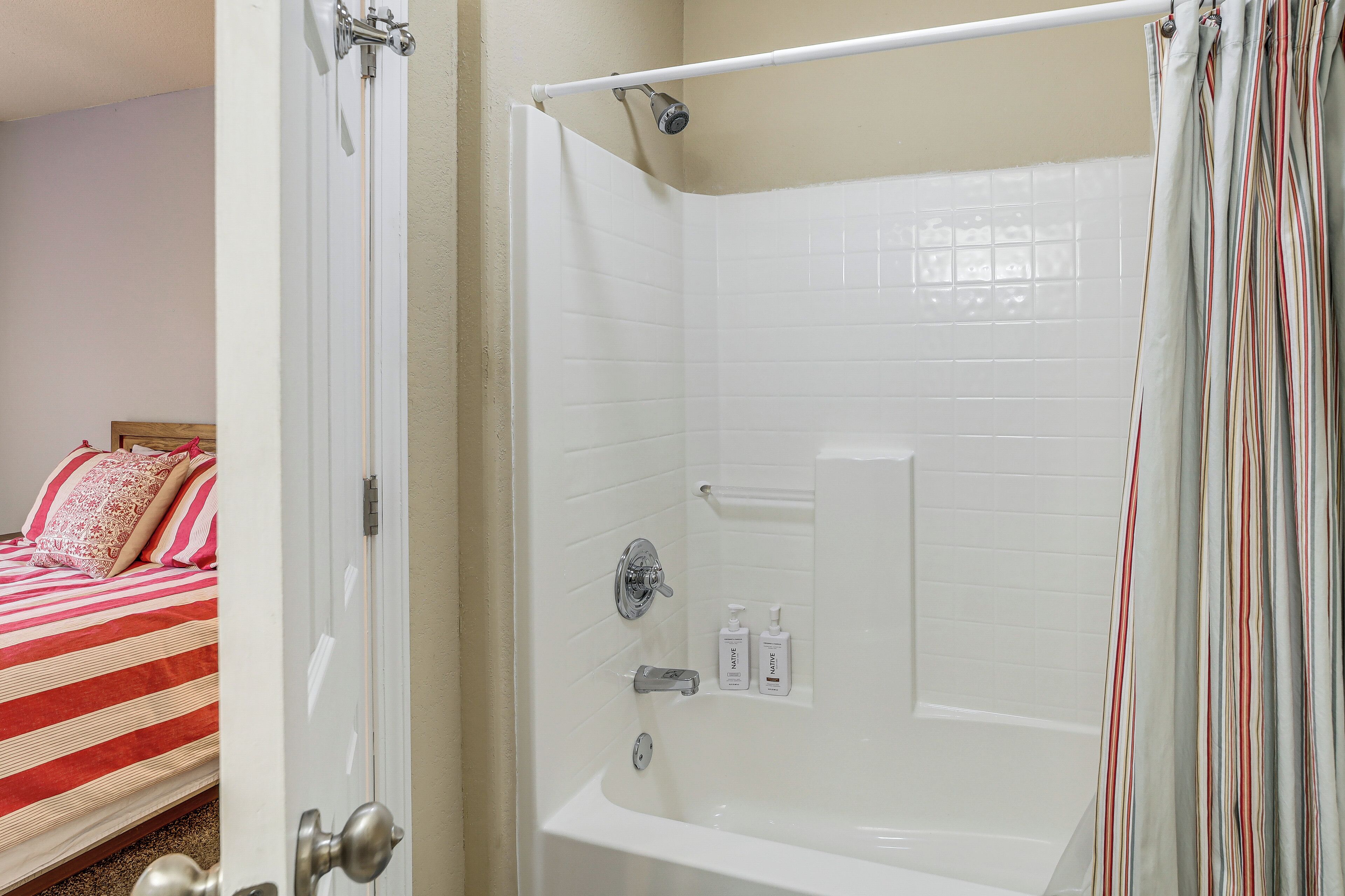 A bathroom with a bathtub, a shower, and a toilet. There is a bed with striped bedding in the adjacent room.