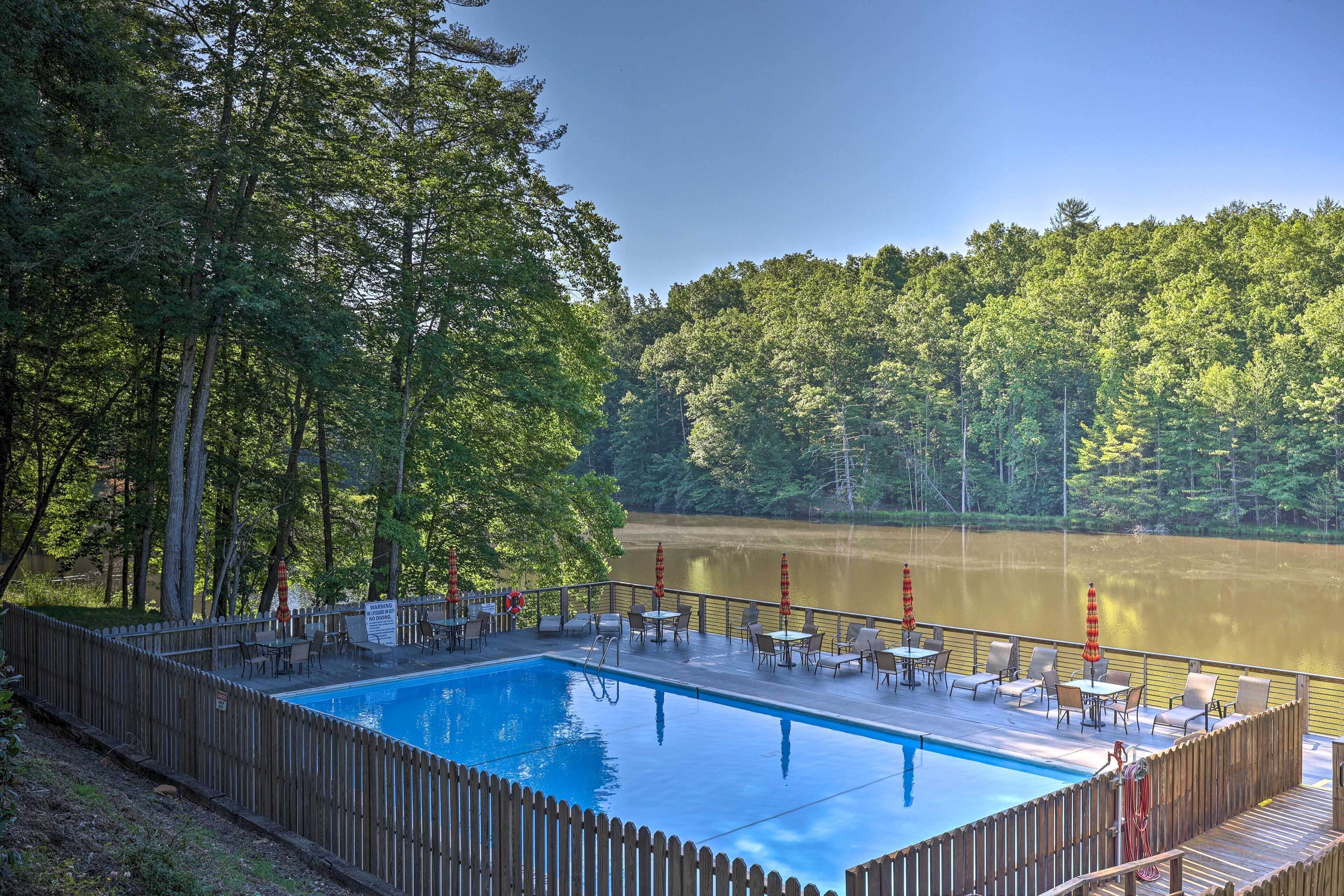 A rectangular pool with a wooden deck and chairs, surrounded by a wooden fence, overlooking a lake with trees in the background.