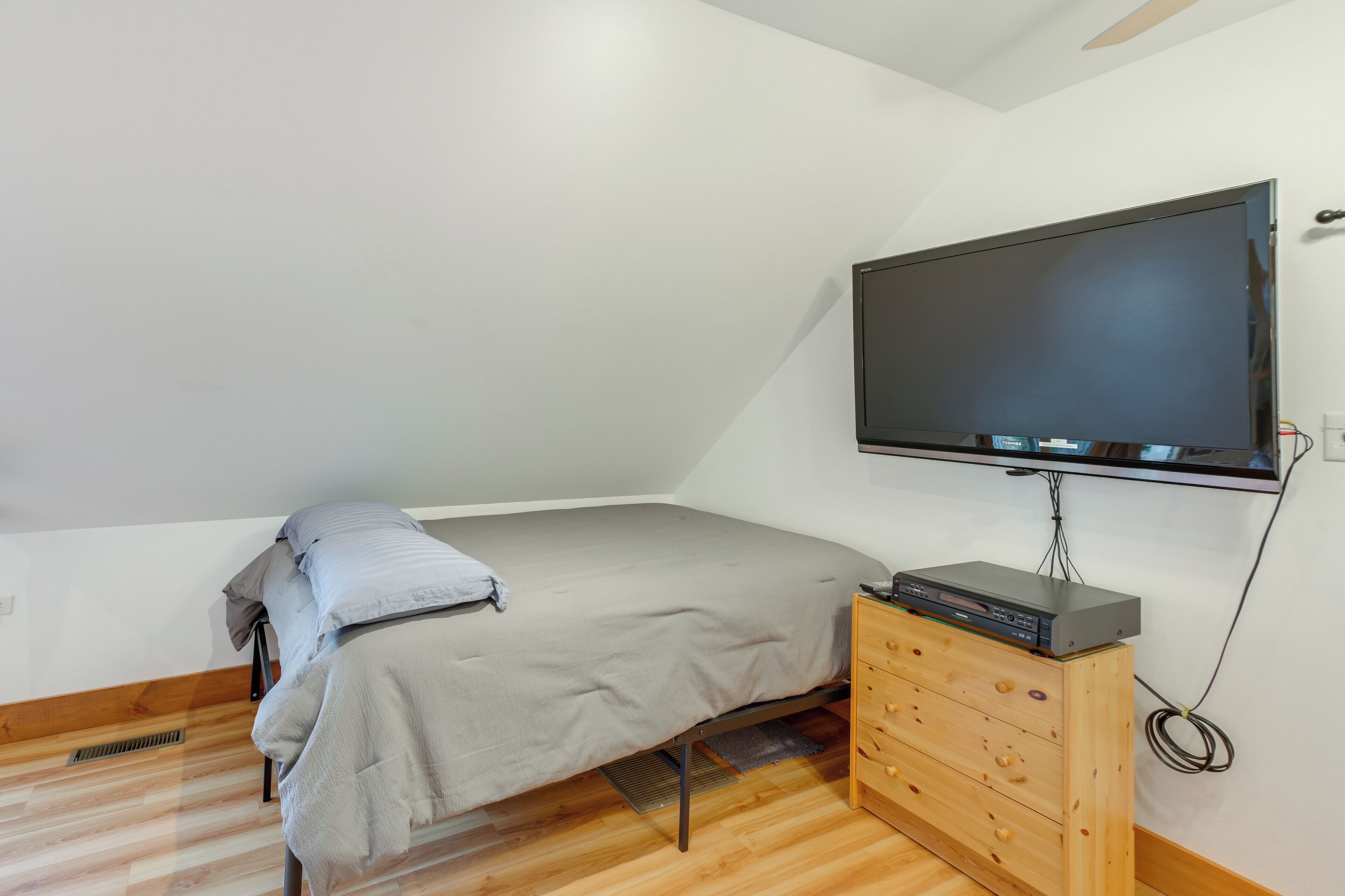 A small, single-bed room with a television mounted on the wall, a wooden dresser, and a vent on the floor.