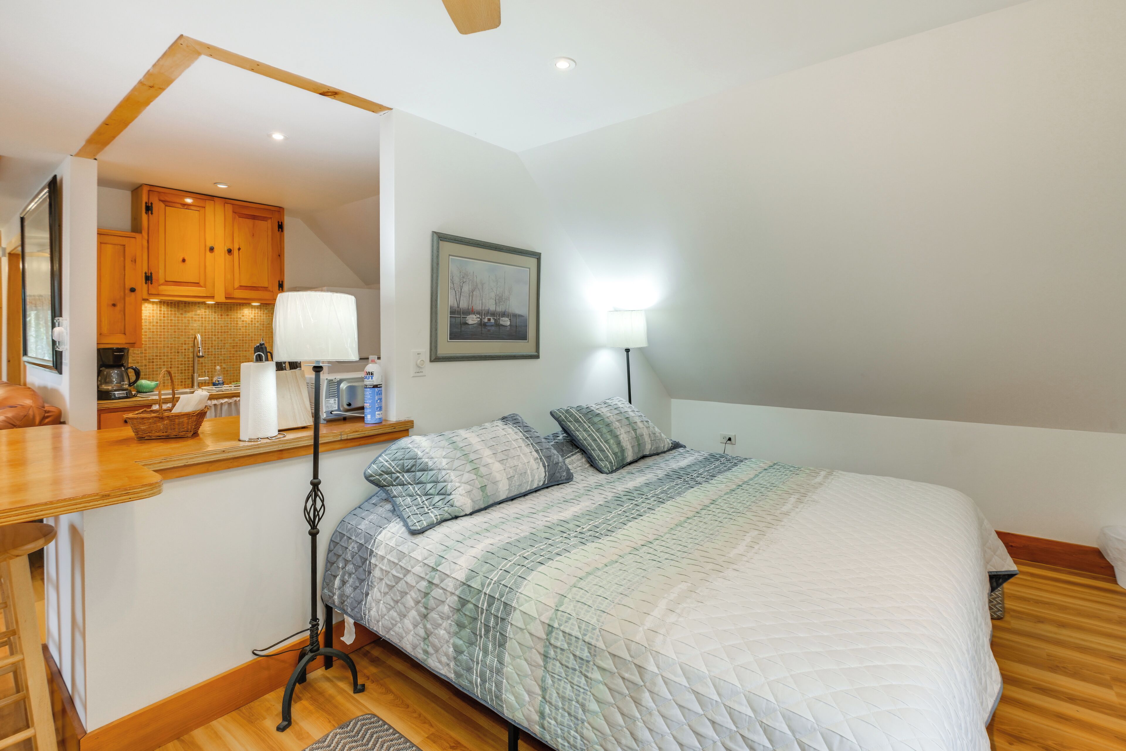 A bedroom with a bed, bedside table, lamp, and a view of the kitchen.