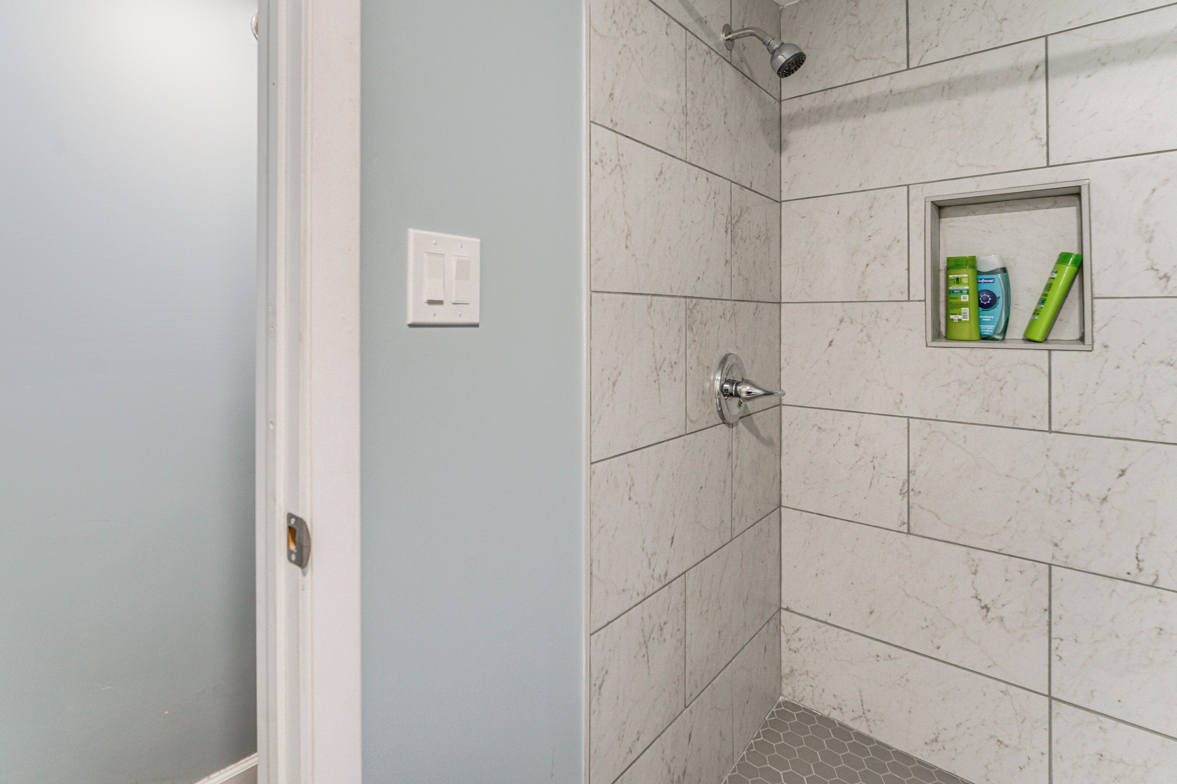 A bathroom with white marble tiles, a shower with a handheld showerhead, and a small shelf with toiletries.