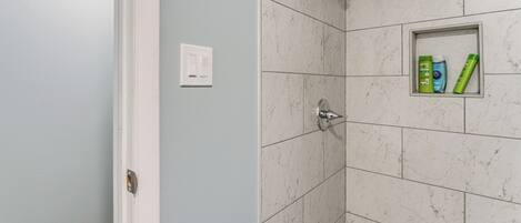 A bathroom with white marble tiles, a shower with a handheld showerhead, and a small shelf with toiletries.