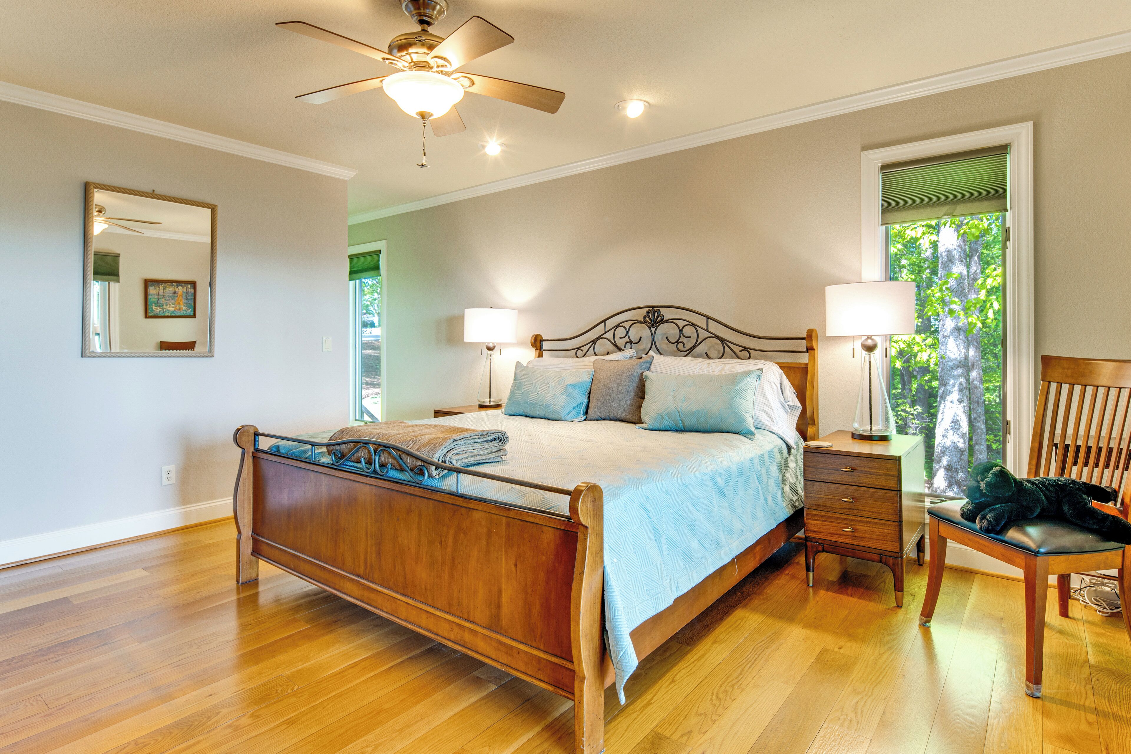 A bedroom with a wooden bed, two bedside tables with lamps, a ceiling fan, and a window with a view of trees.