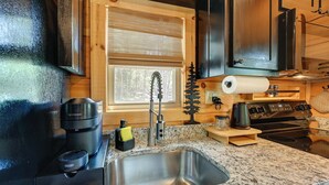 A kitchen with a stainless steel sink, a coffee maker, a stove, and a window with a view of trees.