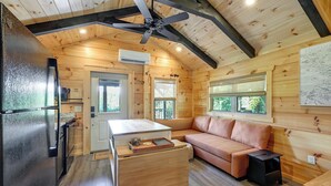 A wooden cabin interior with a kitchenette, a sofa, a ceiling fan, and a window with a view of greenery.